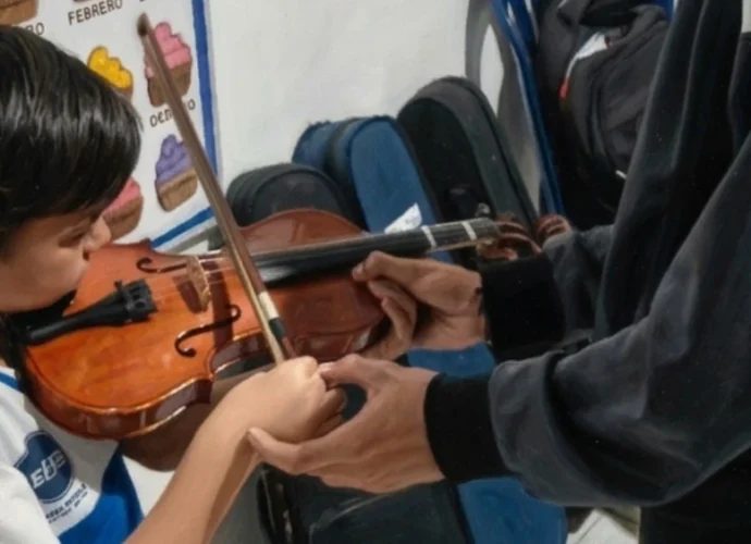 Un niño estudiante de la U.E. Johnny Escobar sosteniendo un violín durante una clase de iniciación musical, mientras un instructor guía la posición de sus manos.