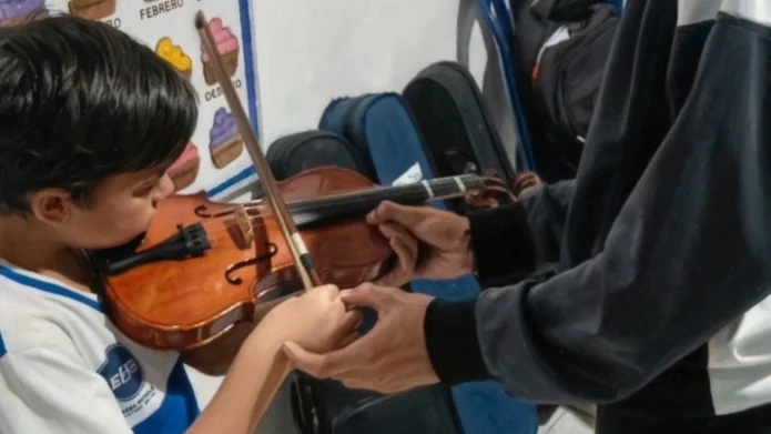 Un niño estudiante de la U.E. Johnny Escobar sosteniendo un violín durante una clase de iniciación musical, mientras un instructor guía la posición de sus manos.