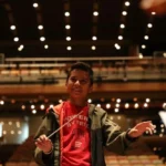 Primer plano de Samuel Abraham González Quintero sonriendo y dirigiendo con batuta en la Sala Simón Bolívar de Caracas, ilustrando el éxito del plan de carrera de Otilca.