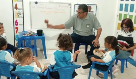Maestro Eliut González en un salón de clases interactúa con un grupo de niños de preescolar sentados en círculo, mostrándoles una maraca, mientras la maestra Liliana Rivas está sentada a un lado revisando un cuaderno.
