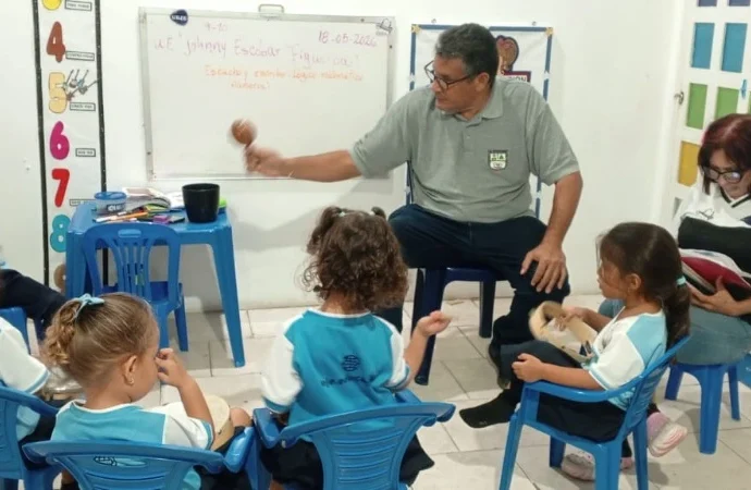 Maestro Eliut González en un salón de clases interactúa con un grupo de niños de preescolar sentados en círculo, mostrándoles una maraca, mientras la maestra Liliana Rivas está sentada a un lado revisando un cuaderno.