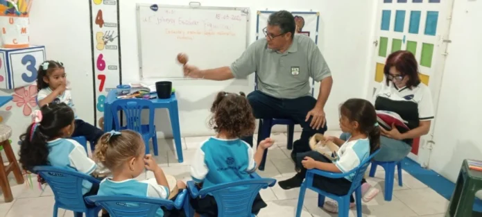 Maestro Eliut González en un salón de clases interactúa con un grupo de niños de preescolar sentados en círculo, mostrándoles una maraca, mientras la maestra Liliana Rivas está sentada a un lado revisando un cuaderno.
