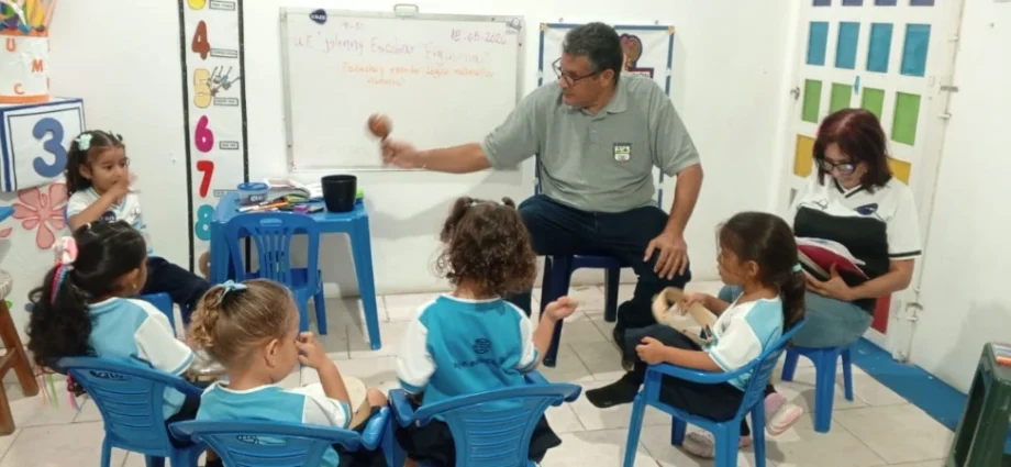 Maestro Eliut González en un salón de clases interactúa con un grupo de niños de preescolar sentados en círculo, mostrándoles una maraca, mientras la maestra Liliana Rivas está sentada a un lado revisando un cuaderno.